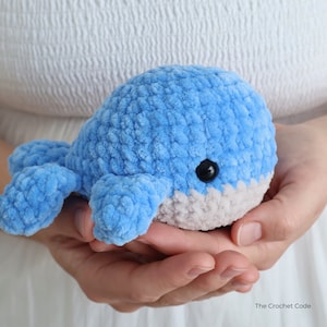 Op de afbeelding: Een handgemaakte, blauwe gehaakte walvis met een grijze buik en zwarte ogen. Het pluche speelgoed wordt in open handen gehouden, waarbij de zachte textuur en ronde vorm worden getoond. Het logo The Crochet Code is zichtbaar.