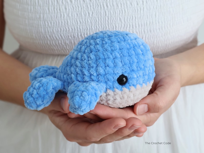 Op de afbeelding: Een handgemaakte, blauwe gehaakte walvis met een grijze buik en zwarte ogen. Het pluche speelgoed wordt in open handen gehouden, waarbij de zachte textuur en ronde vorm worden getoond. Het logo The Crochet Code is zichtbaar.