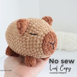 May include: A crocheted capybara plushie in shades of tan and brown. The handmade stuffed animal has a rounded body, small ears, and closed eyes. The text "No sew Loaf Capy Crochet Pattern" is visible.