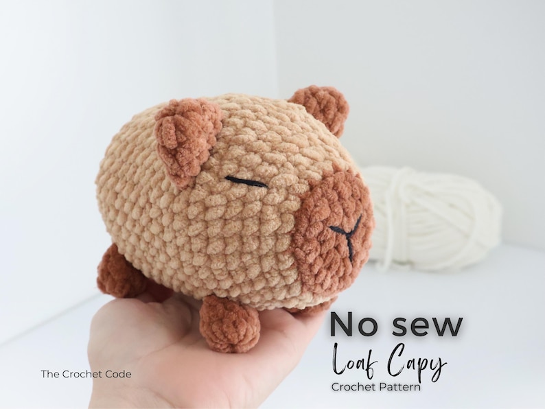 May include: A crocheted capybara plushie in shades of tan and brown. The handmade stuffed animal has a rounded body, small ears, and closed eyes. The text "No sew Loaf Capy Crochet Pattern" is visible.