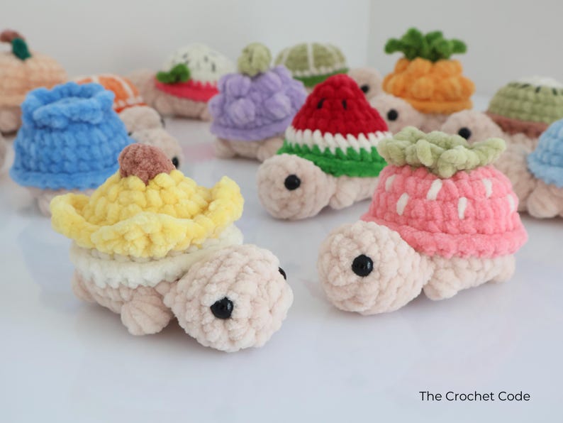 May include: A collection of crocheted turtle toys, each with a different fruit-themed shell. The turtles are in various colors, including pink, yellow, blue, and green. The toys have black button eyes and are displayed on a white surface.