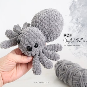 Pictured against a ball of yarn is a fog grey crochet spider. The zoomed in detail shot of the DIY amigurumi features four eyes staring directly at the viewer.