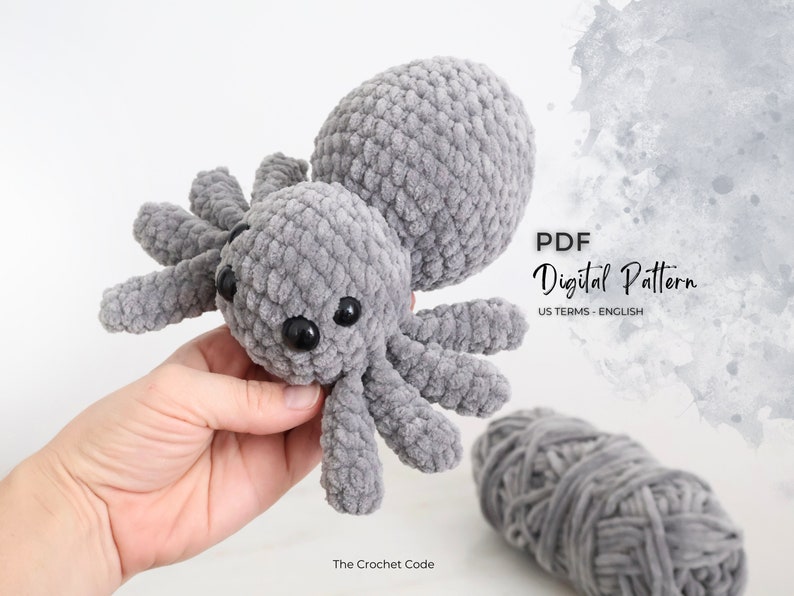 Pictured against a ball of yarn is a fog grey crochet spider. The zoomed in detail shot of the DIY amigurumi features four eyes staring directly at the viewer.