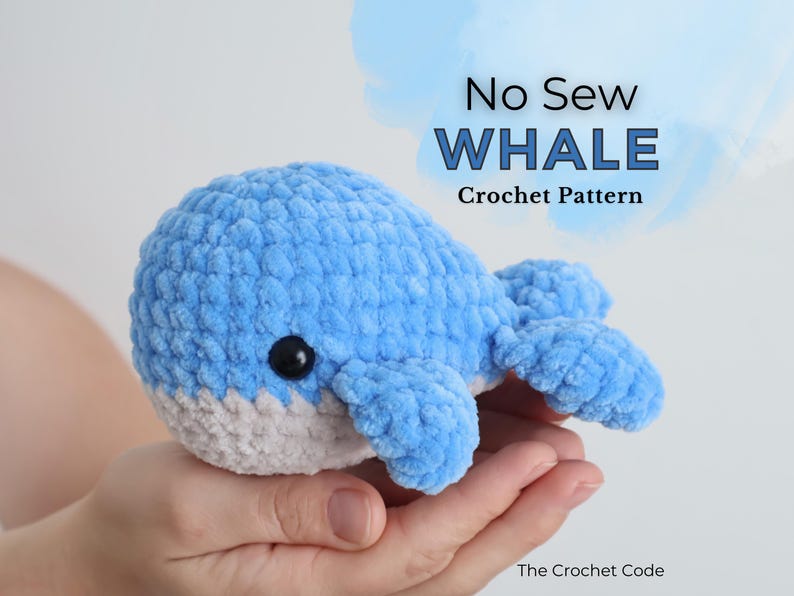 Op de afbeelding: Een handgemaakte, blauw-witte gehaakte walvis met een zwart oog. Het pluche speelgoed wordt in de handen van een persoon gehouden. Tekst op de afbeelding luidt "No Sew WHALE Crochet Pattern" en "The Crochet Code".