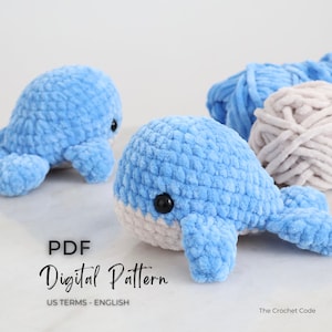 Op de afbeelding: Twee gehaakte walvissen in lichtblauw met witte buiken en zwarte ogen worden getoond. Daarnaast liggen twee strengen blauw garen en een streng beige garen. De tekst "PDF Digital Pattern US Terms - English" is ook zichtbaar.