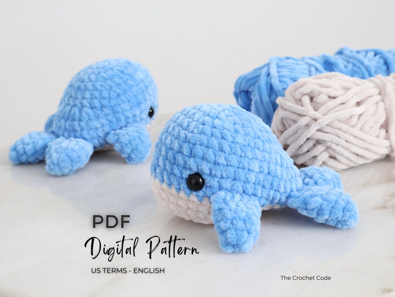 Op de afbeelding: Twee gehaakte walvissen in lichtblauw met witte buiken en zwarte ogen worden getoond. Daarnaast liggen twee strengen blauw garen en een streng beige garen. De tekst "PDF Digital Pattern US Terms - English" is ook zichtbaar.