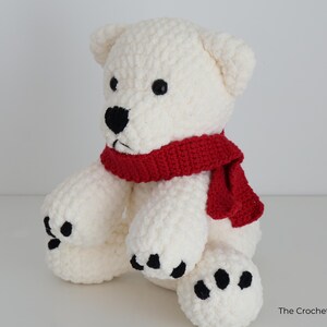 May include: A handmade, white crochet bear with black eyes and nose, and black paw pads. The bear wears a red scarf around its neck. The Crochet Code is printed on the bottom right corner.