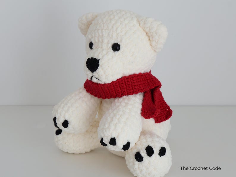 May include: A handmade, white crochet bear with black eyes and nose, and black paw pads. The bear wears a red scarf around its neck. The Crochet Code is printed on the bottom right corner.