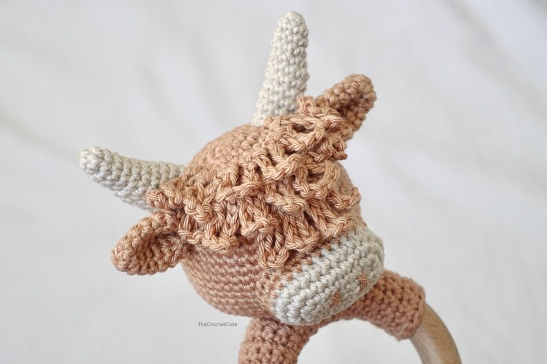 May include: A crocheted cow rattle toy in brown and white. The toy has a wooden ring attached to the bottom.