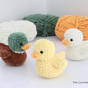 May include: Three crocheted ducklings in brown, yellow, and white, each with an orange beak and black eyes. The ducks are in front of three balls of yarn in white, green, and orange. The Crochet Code.