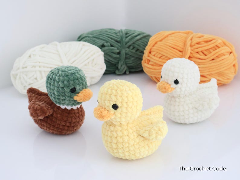 May include: Three crocheted ducklings in brown, yellow, and white, each with an orange beak and black eyes. The ducks are in front of three balls of yarn in white, green, and orange. The Crochet Code.