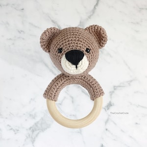 May include: A brown crocheted bear head attached to a wooden teething ring. The bear has black eyes and a black nose.
