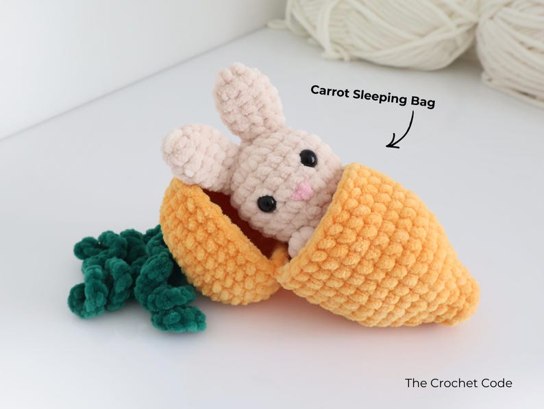 May include: A crocheted bunny peeks out of an orange carrot sleeping bag, with green leaves at the base. The bunny is light pink with black eyes and a pink nose. The text "Carrot Sleeping Bag" is above the carrot.