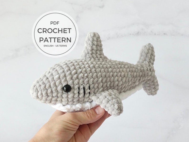 May include: A gray crocheted shark plush toy with black eyes and a white belly. The toy is being held in a hand. The image includes the text "PDF CROCHET PATTERN ENGLISH - US TERMS".