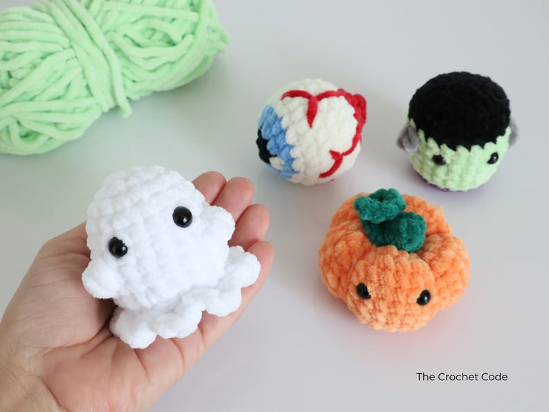 May include: A selection of handmade crochet Halloween-themed plush toys. Includes a white ghost, an eyeball, a Frankenstein's monster, and an orange pumpkin with a green stem. The Crochet Code is visible in the bottom right corner.