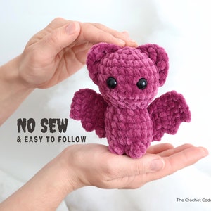 May include: A pink crocheted bat with black eyes. The bat is being held in the hands of a person. The text "NO SEW & EASY TO FOLLOW" is visible below the bat.