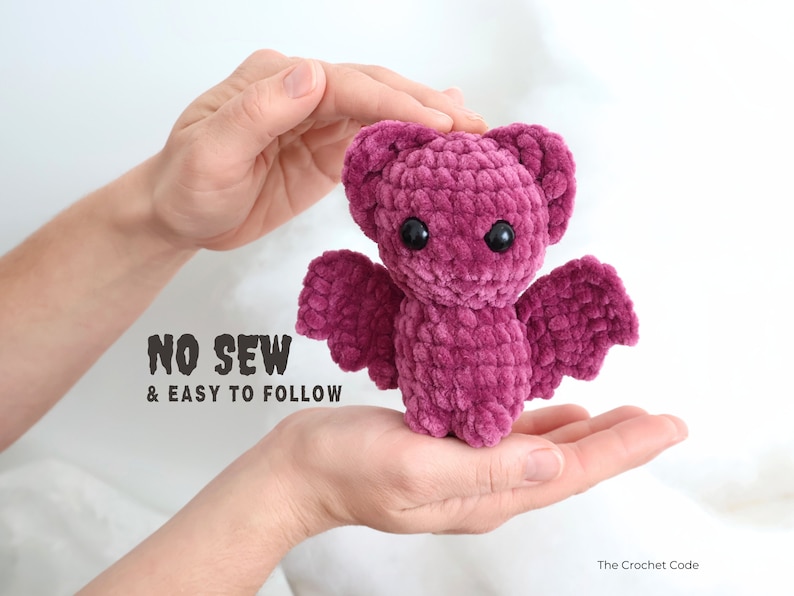 May include: A pink crocheted bat with black eyes. The bat is being held in the hands of a person. The text "NO SEW & EASY TO FOLLOW" is visible below the bat.