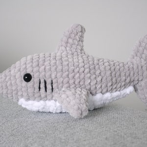 May include: A gray crocheted shark plush toy with black eyes and a white belly. The shark is lying on a gray textured surface.
