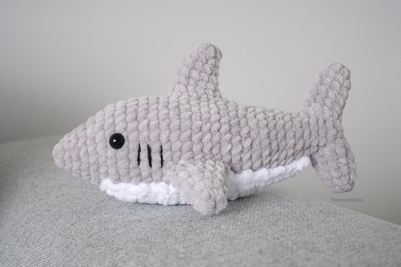 May include: A gray crocheted shark plush toy with black eyes and a white belly. The shark is lying on a gray textured surface.