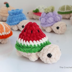 May include: A collection of handmade crochet turtles, each with a different fruit-themed shell. The turtles feature a variety of colors, including red, green, white, orange, and purple. The turtles are made of soft yarn and have black eyes.