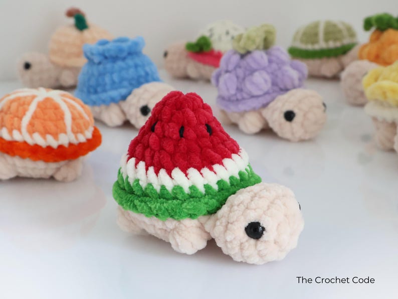 May include: A collection of handmade crochet turtles, each with a different fruit-themed shell. The turtles feature a variety of colors, including red, green, white, orange, and purple. The turtles are made of soft yarn and have black eyes.