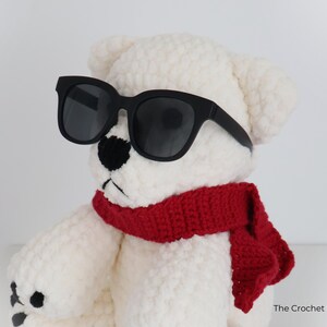 May include: A white crocheted teddy bear wearing black sunglasses and a red scarf. The bear has a black nose and black stitched details. The Crochet Code is printed on the bottom right.