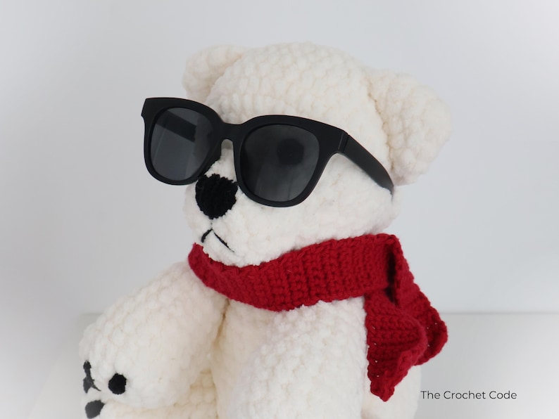 May include: A white crocheted teddy bear wearing black sunglasses and a red scarf. The bear has a black nose and black stitched details. The Crochet Code is printed on the bottom right.