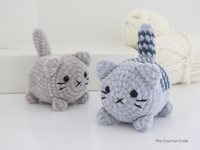 May include: Two handmade crochet cat toys. One is solid gray, the other has a blue and gray striped tail and back. Both have black eyes and whiskers. The toys are small and round, and appear to be made of soft yarn.