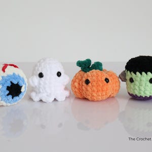May include: Four crocheted Halloween figures: an eyeball, a ghost, a pumpkin, and Frankenstein's monster. The eyeball is white, blue, and red. The ghost is white. The pumpkin is orange. The monster is green. The Crochet Code is written on the bottom right.