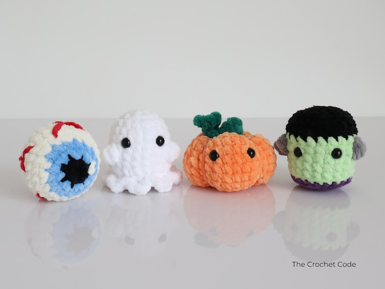 May include: Four crocheted Halloween figures: an eyeball, a ghost, a pumpkin, and Frankenstein's monster. The eyeball is white, blue, and red. The ghost is white. The pumpkin is orange. The monster is green. The Crochet Code is written on the bottom right.