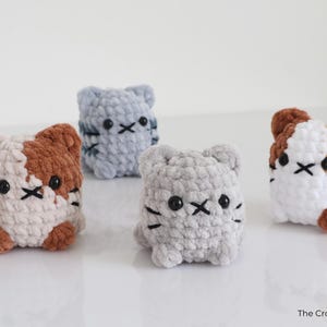 May include: Four handmade crochet cat figures in various colors: gray, light blue, white with brown, and white with orange. Each cat has black eyes and a stitched mouth. The Crochet Code is in the bottom right corner.