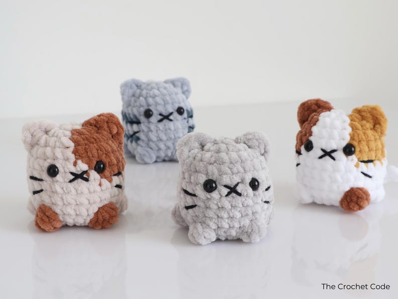 May include: Four handmade crochet cat figures in various colors: gray, light blue, white with brown, and white with orange. Each cat has black eyes and a stitched mouth. The Crochet Code is in the bottom right corner.