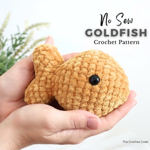 May include: A crocheted goldfish toy in a light brown colour with black eyes. The toy is being held in a person's hands. The text "No Sew GOLDFISH Crochet Pattern" is visible above the toy.