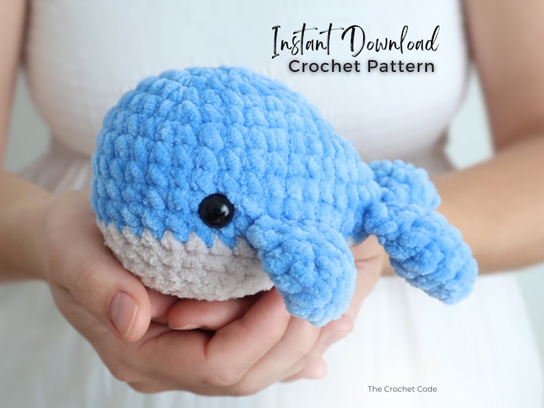 Op de afbeelding: Een handgemaakte, lichtblauwe gehaakte walvis met een grijze buik en zwarte knoopogen. De zachte, pluche walvis wordt in open handen gehouden. De afbeelding bevat de tekst "Instant Download Crochet Pattern".