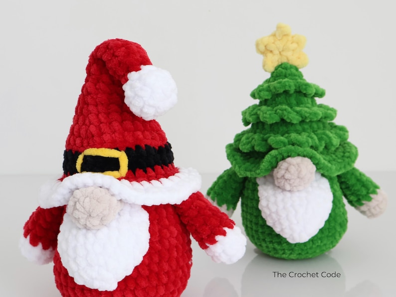 Puede incluir: Dos gnomos navide&ntilde;os de peluche. Uno es rojo con barba y ribete de sombrero blancos, cintur&oacute;n negro y hebilla dorada. El otro es un gnomo de &aacute;rbol de Navidad verde con una estrella amarilla y barba blanca. The Crochet Code.