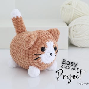 May include: A handmade crochet cat in shades of brown and white, with black facial details. The cat has a fluffy texture and a small tail. The image includes the text "Easy Crochet Project" and two balls of white yarn.