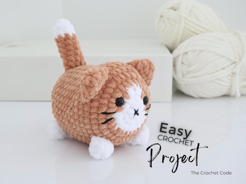 May include: A handmade crochet cat in shades of brown and white, with black facial details. The cat has a fluffy texture and a small tail. The image includes the text "Easy Crochet Project" and two balls of white yarn.