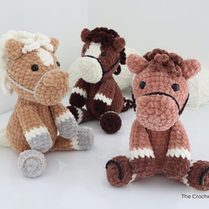 Three handmade crochet horse plushies in shades of brown, tan, and white. Each plush horse has a unique color pattern, with details like manes, hooves, and facial features. The horses are sitting on a white surface.