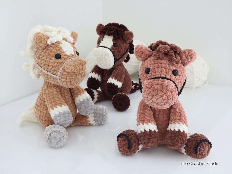 Three handmade crochet horse plushies in shades of brown, tan, and white. Each plush horse has a unique color pattern, with details like manes, hooves, and facial features. The horses are sitting on a white surface.
