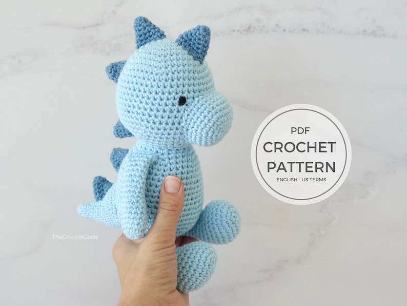 May include: A light blue crocheted dinosaur amigurumi toy with black eyes. The toy is being held by a hand. The image includes the text "PDF CROCHET PATTERN ENGLISH - US TERMS".