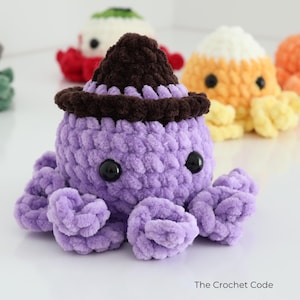 Puede incluir: Un peluche de pulpo tejido a ganchillo de color morado con un sombrero de bruja marr&oacute;n oscuro y ojos de pl&aacute;stico negro. El peluche tiene una superficie de hilo texturizado y est&aacute; rodeado de otras criaturas tejidas. El texto The Crochet Code es visible.
