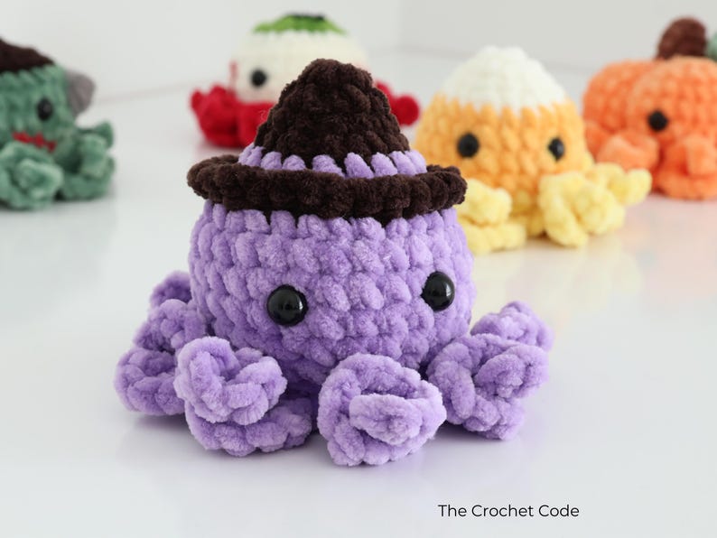 Puede incluir: Un peluche de pulpo tejido a ganchillo de color morado con un sombrero de bruja marr&oacute;n oscuro y ojos de pl&aacute;stico negro. El peluche tiene una superficie de hilo texturizado y est&aacute; rodeado de otras criaturas tejidas. El texto The Crochet Code es visible.