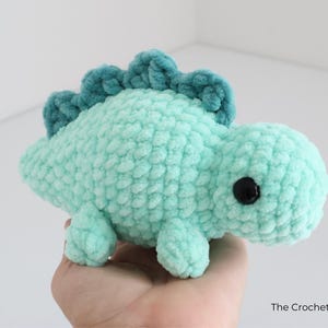 May include: A handmade, turquoise crochet dinosaur plushie with a textured, fuzzy appearance. The dinosaur has a darker teal ridge along its back and black safety eyes. The Crochet Code is written on the bottom right.
