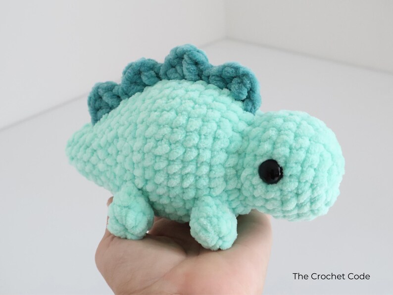 May include: A handmade, turquoise crochet dinosaur plushie with a textured, fuzzy appearance. The dinosaur has a darker teal ridge along its back and black safety eyes. The Crochet Code is written on the bottom right.