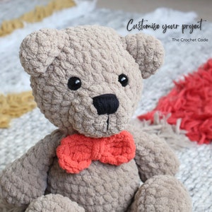 May include: A crocheted teddy bear with a gray fur texture and a red bow tie. The bear is sitting on a white and red rug. The text "Customise your project" is visible in the image.