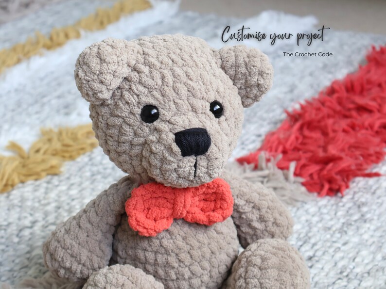 May include: A crocheted teddy bear with a gray fur texture and a red bow tie. The bear is sitting on a white and red rug. The text "Customise your project" is visible in the image.