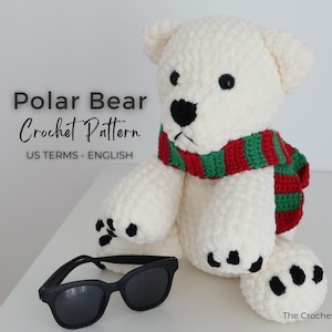 May include: A white crochet polar bear wearing a red and green striped scarf, with black sunglasses resting in front. The bear has black eyes and paw pads. Text on the image reads "Polar Bear Crochet Pattern US Terms - English".