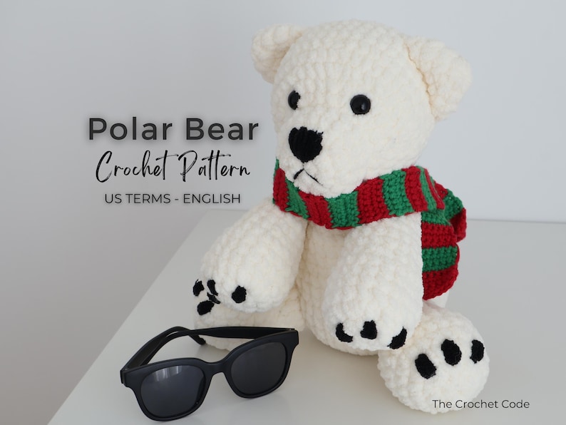 May include: A white crochet polar bear wearing a red and green striped scarf, with black sunglasses resting in front. The bear has black eyes and paw pads. Text on the image reads "Polar Bear Crochet Pattern US Terms - English".