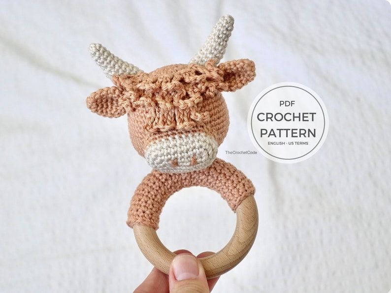 May include: Crochet pattern for a brown and white cow rattle toy with a wooden ring. The pattern is in English and uses US terms. The pattern is available as a PDF download.