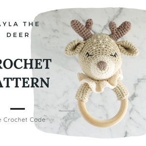 Peut inclure: Un hochet en forme de cerf crocheté avec une base en anneau de bois. Le cerf a des accents beiges et marron, avec le texte "AYLA THE DEER CROCHET PATTERN" et "The Crochet Code" en noir. Le hochet est sur un fond de marbre blanc.